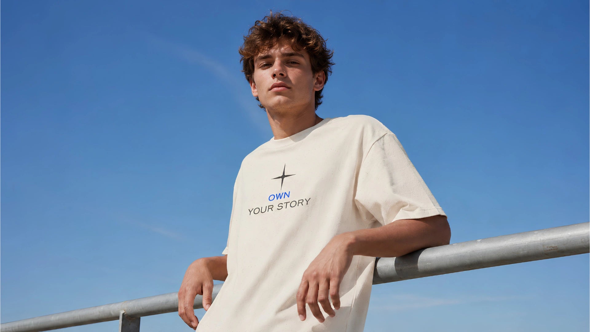Young man wearing a cream Stillor Clothing “Own Your Story” T-shirt, leaning against a guardrail under a clear blue sky.