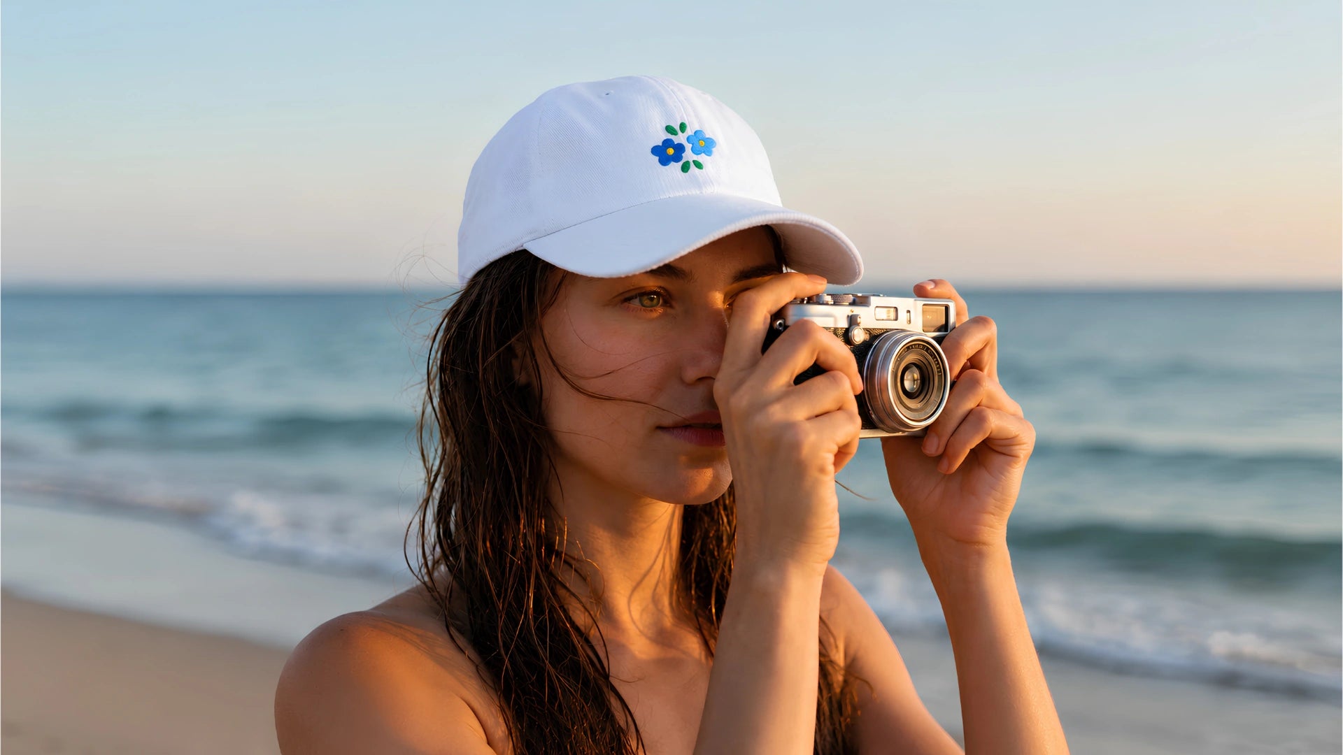 Woman wearing a white Stillor Clothing embroidered flower cap, holding a vintage camera at the beach during golden hour.